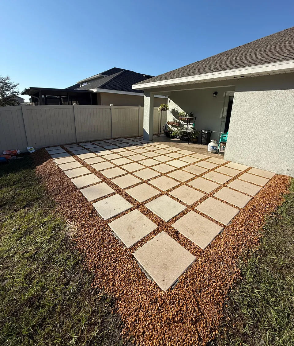 Side-yard patio of large square concrete pavers on a gravel bed with open gravel joints, beside a single-story home and vinyl fence