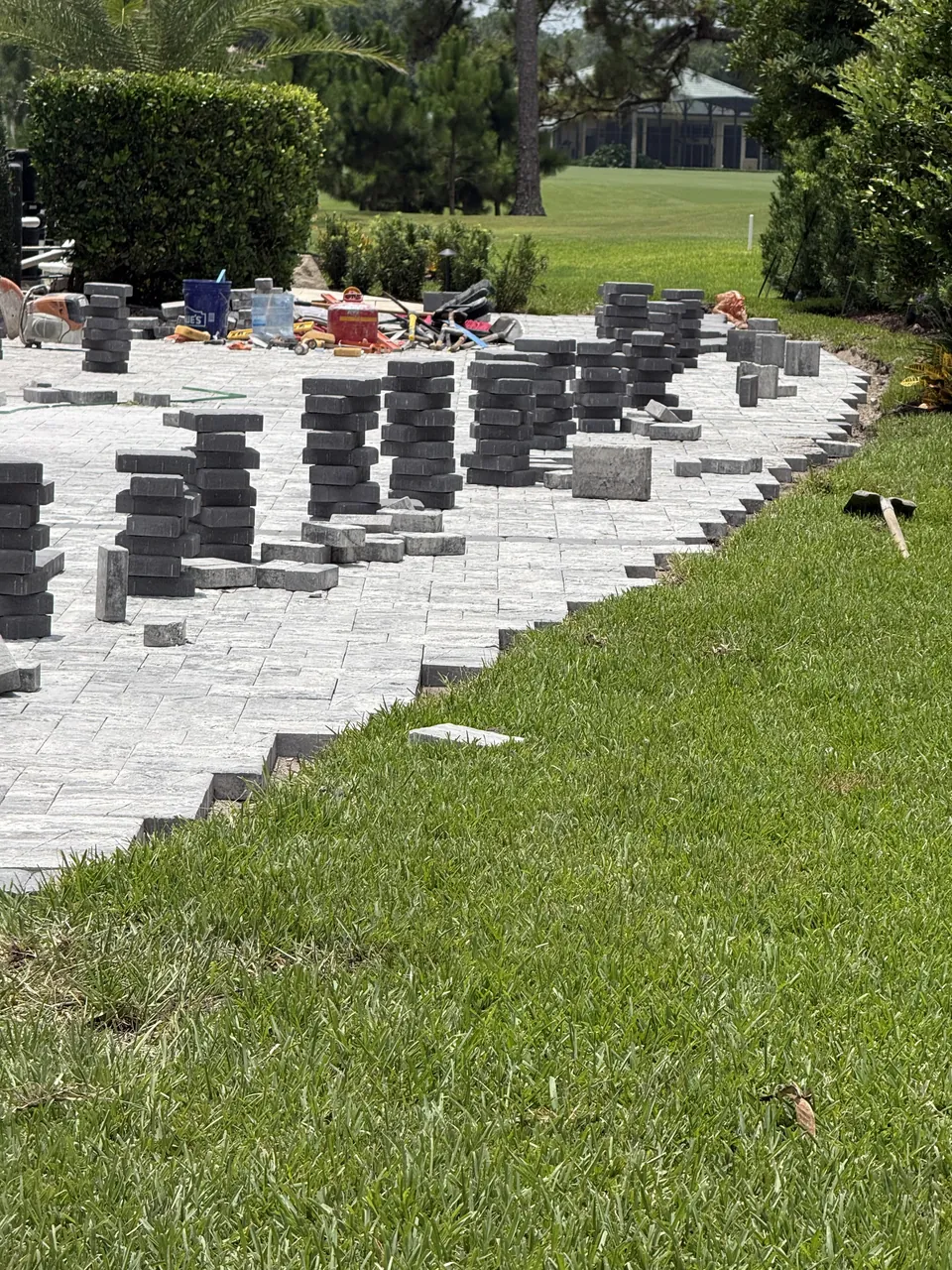Stacked pavers staged along a driveway during installation, showing the leveled bedding and edge line against the lawn
