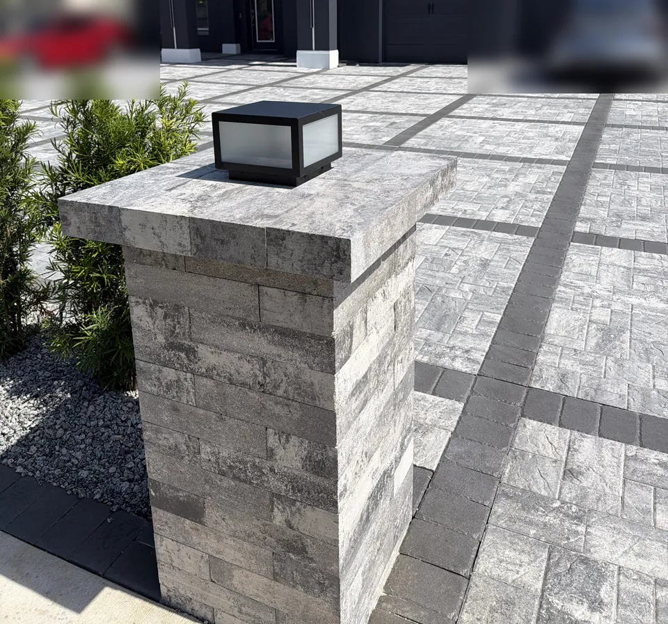 Paver-clad driveway entry pillar with a wall-mounted light, matching the gray and charcoal paver driveway behind it