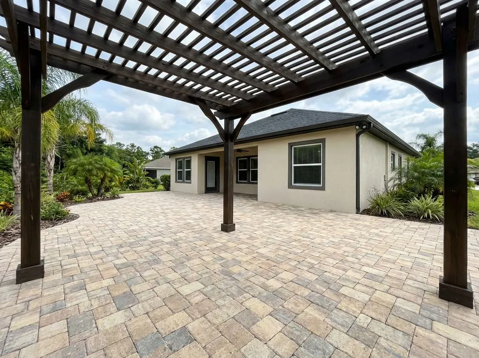 Large paver patio in a tumbled tan blend under a dark-stained wood pergola behind a single-story home