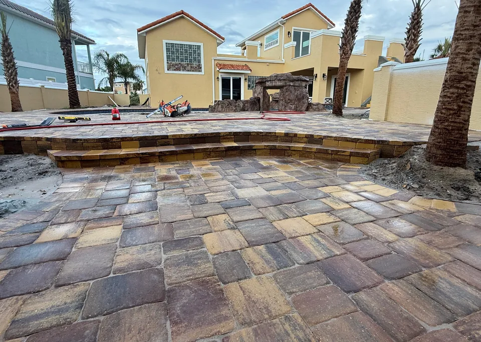 Multi-level paver patio under construction with a raised pool terrace, paver-faced step risers and a wet-cut saw on site